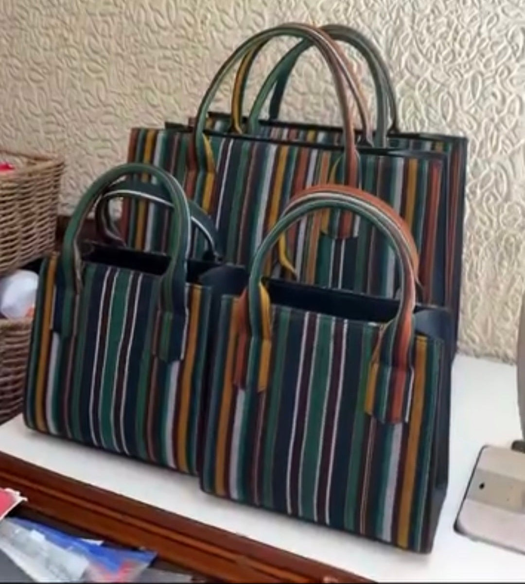 Three striped Aso Oke tote bags in different sizes arranged on a white surface against a textured beige wall.